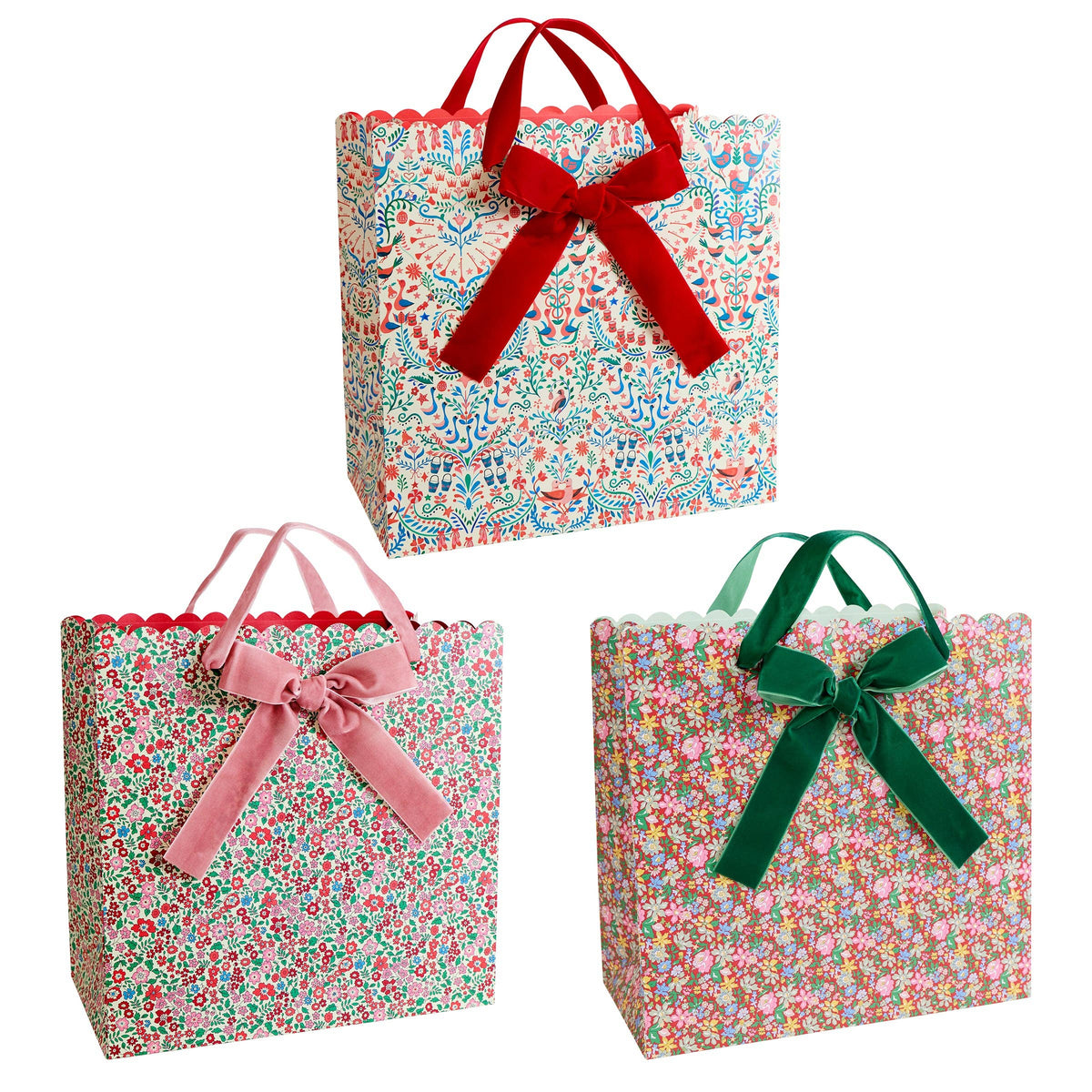 Three floral gift bags arranged on a white background, each with a ribbon handle: one red, one pink, and one green. The bags have a scalloped edge at the top and a dense small floral print.