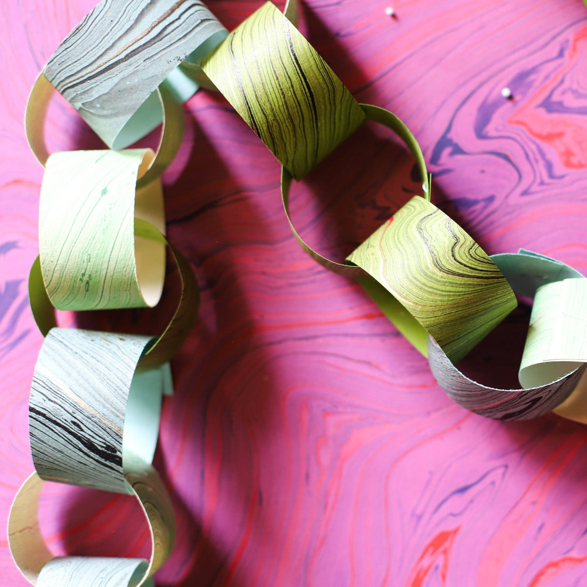 A handmade paper chain garland with alternating marbled paper links in sage green, light blue, and deep gray.