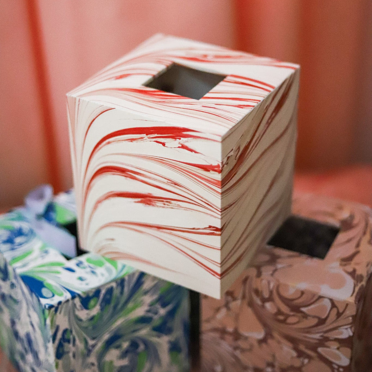 A square paper marbled tissue box cover with a square opening, featuring a white base with red swirls, displayed on top of two other similarly patterned covers.