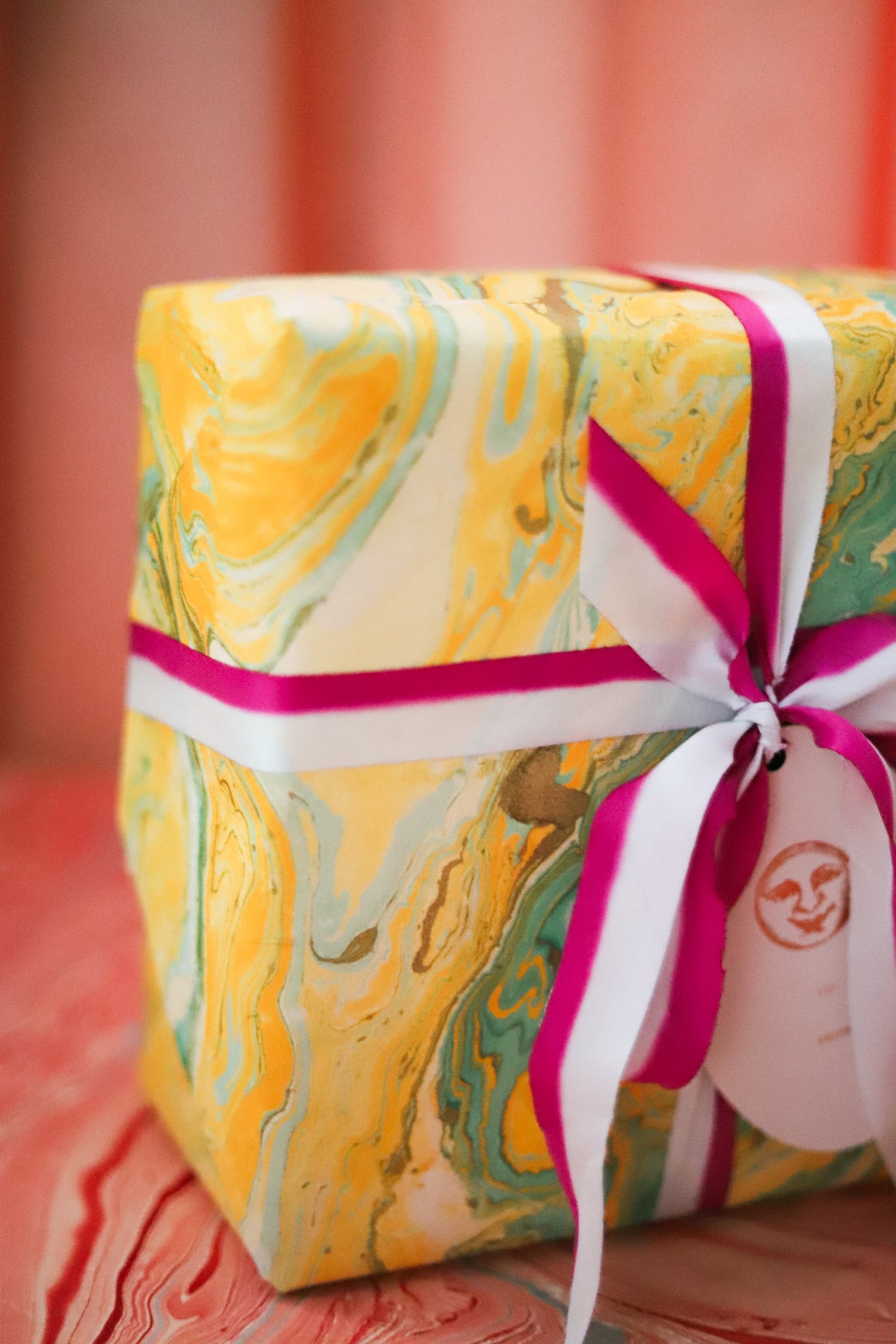 A square gift box wrapped in yellow, green, and white marbled paper with a bright pink ribbon and white bow, tied with a paper tag.