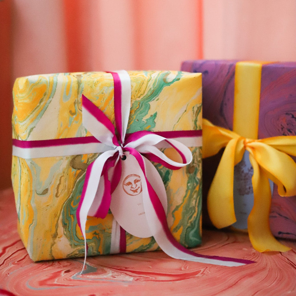 A wrapped gift box in the foreground with yellow, green, and white marbled paper, tied with a pink and white ribbon and a paper tag, next to another wrapped box in purple and yellow wrapping paper against a soft coral background.