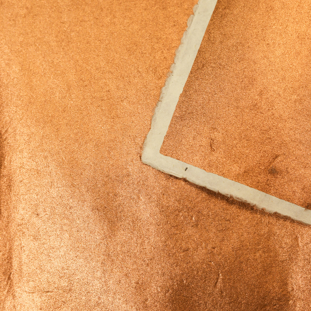 A close-up of a textured metallic copper-orange material with a folded corner showing a light gray backing or edge.