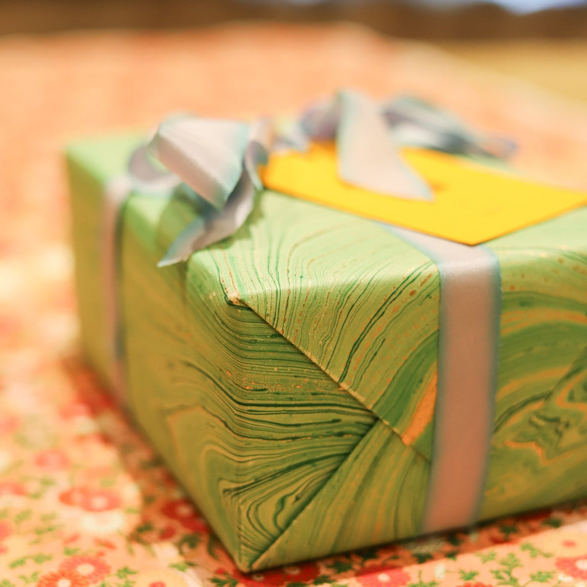A wrapped gift box with green marbled paper, a pale blue ribbon, and a yellow tag, resting on a floral fabric surface.