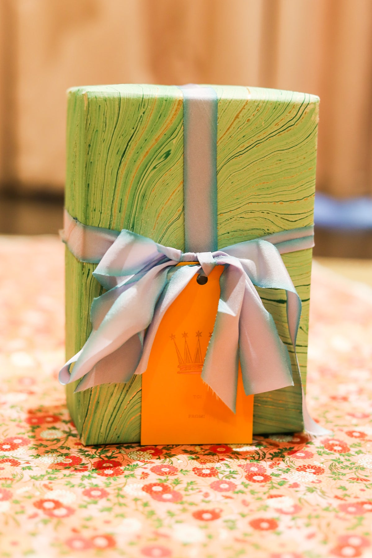 A wrapped gift box with a green marbled pattern, tied with a pale blue ribbon and an orange gift tag, set on a floral surface with a warm blurred background.