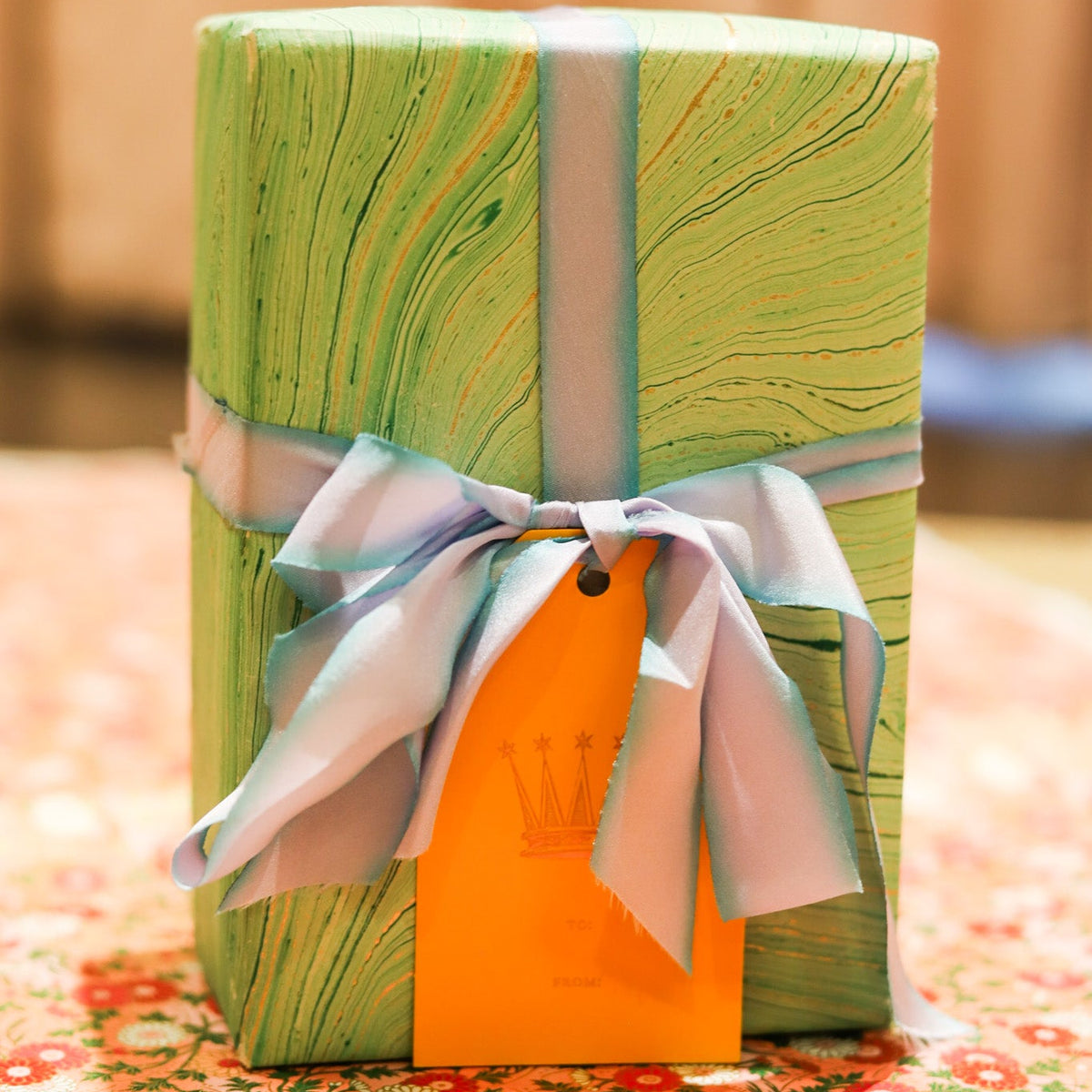 A wrapped gift box with a green marbled pattern, tied with a pale blue ribbon and an orange gift tag, set on a floral surface with a warm blurred background.
