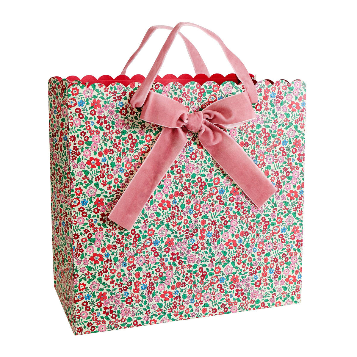 A gift bag with a red, green, and pink floral pattern, featuring a scalloped top edge, pink velvet handles, and a large pink velvet bow on the front.