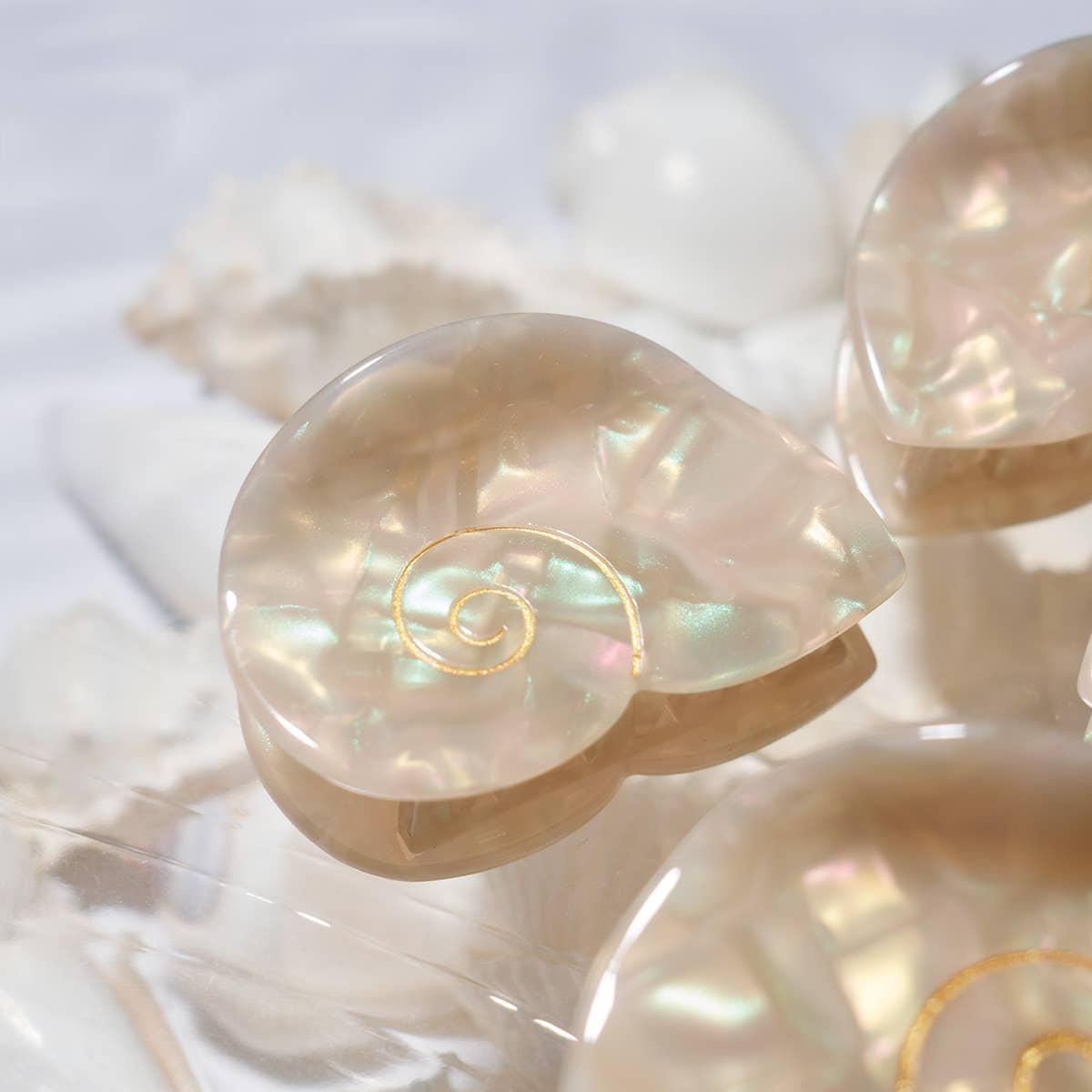 The Siren Hair Claw features a shimmering shell-inspired design with a pearlescent finish and delicate gold spiral detail, crafted from cellulose acetate.