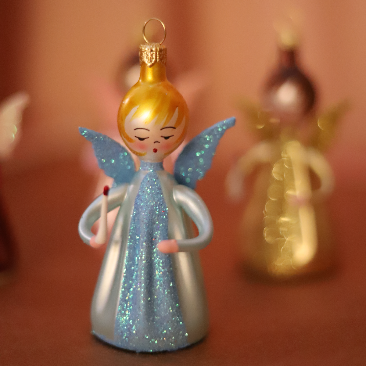 A small angel Christmas ornament with a glittery light blue dress, blue wings, and gold hair, standing on a warm brown surface with a softly blurred second ornament in the background.