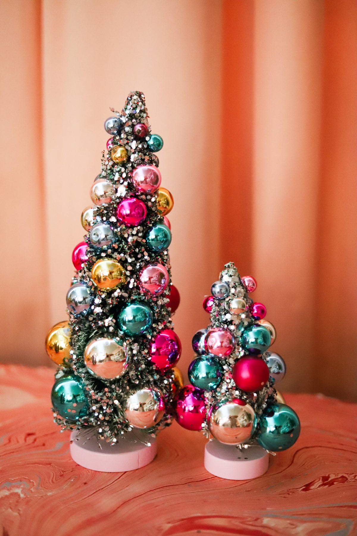Colorful bauble bottle brush Christmas trees with snowy flocking and shimmering ornaments. A festive accent for mantels, villages, tablescapes, and holiday displays.
