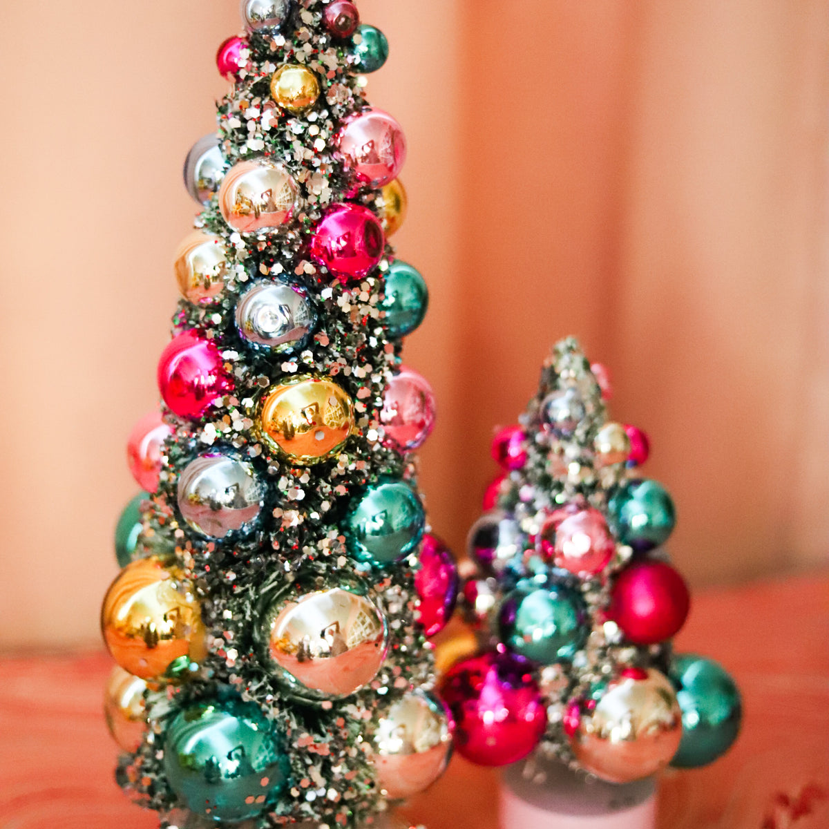 Colorful bauble bottle brush Christmas trees with snowy flocking and shimmering ornaments. A festive accent for mantels, villages, tablescapes, and holiday displays.