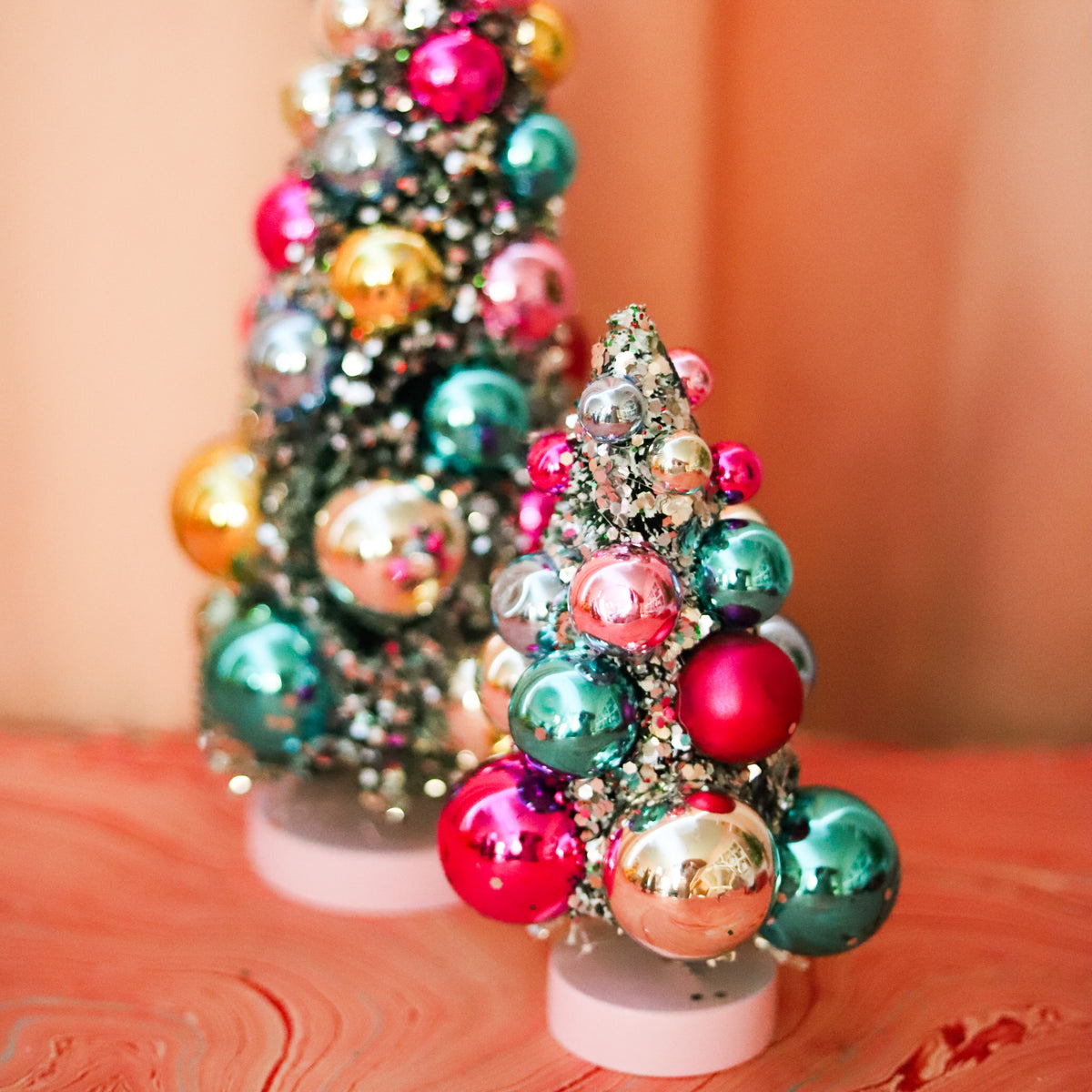 Colorful bauble bottle brush Christmas trees with snowy flocking and shimmering ornaments. A festive accent for mantels, villages, tablescapes, and holiday displays.