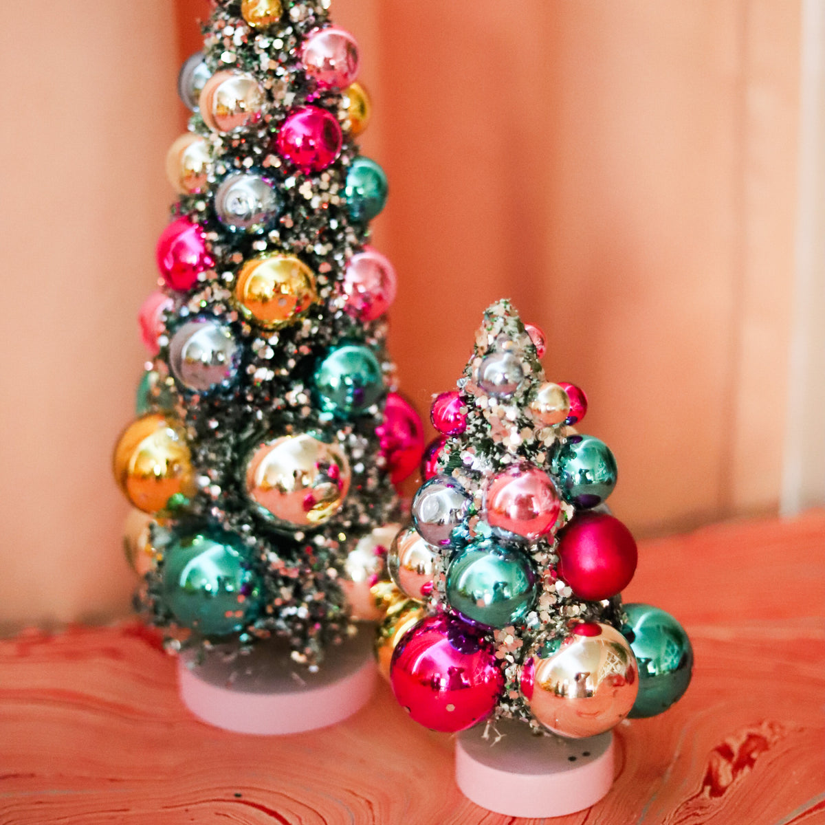 Colorful bauble bottle brush Christmas trees with snowy flocking and shimmering ornaments. A festive accent for mantels, villages, tablescapes, and holiday displays.