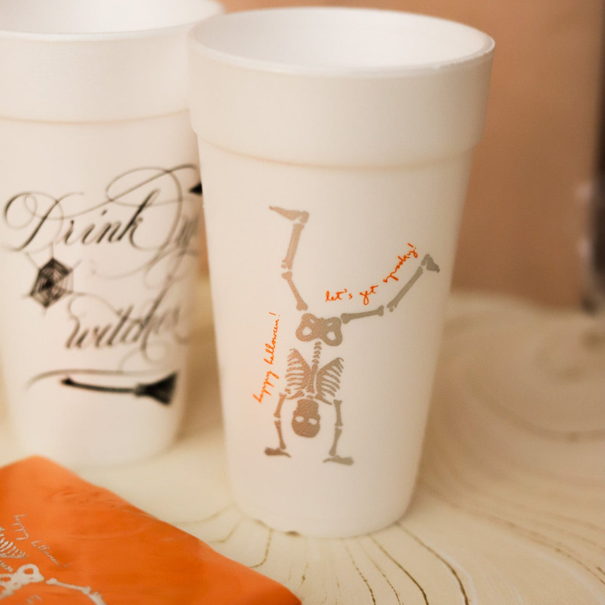 White foam cups with Halloween-themed graphics, including a skeleton handstand and the phrase 'let's go skeleton', displayed on a wooden surface with an orange napkin in the foreground.