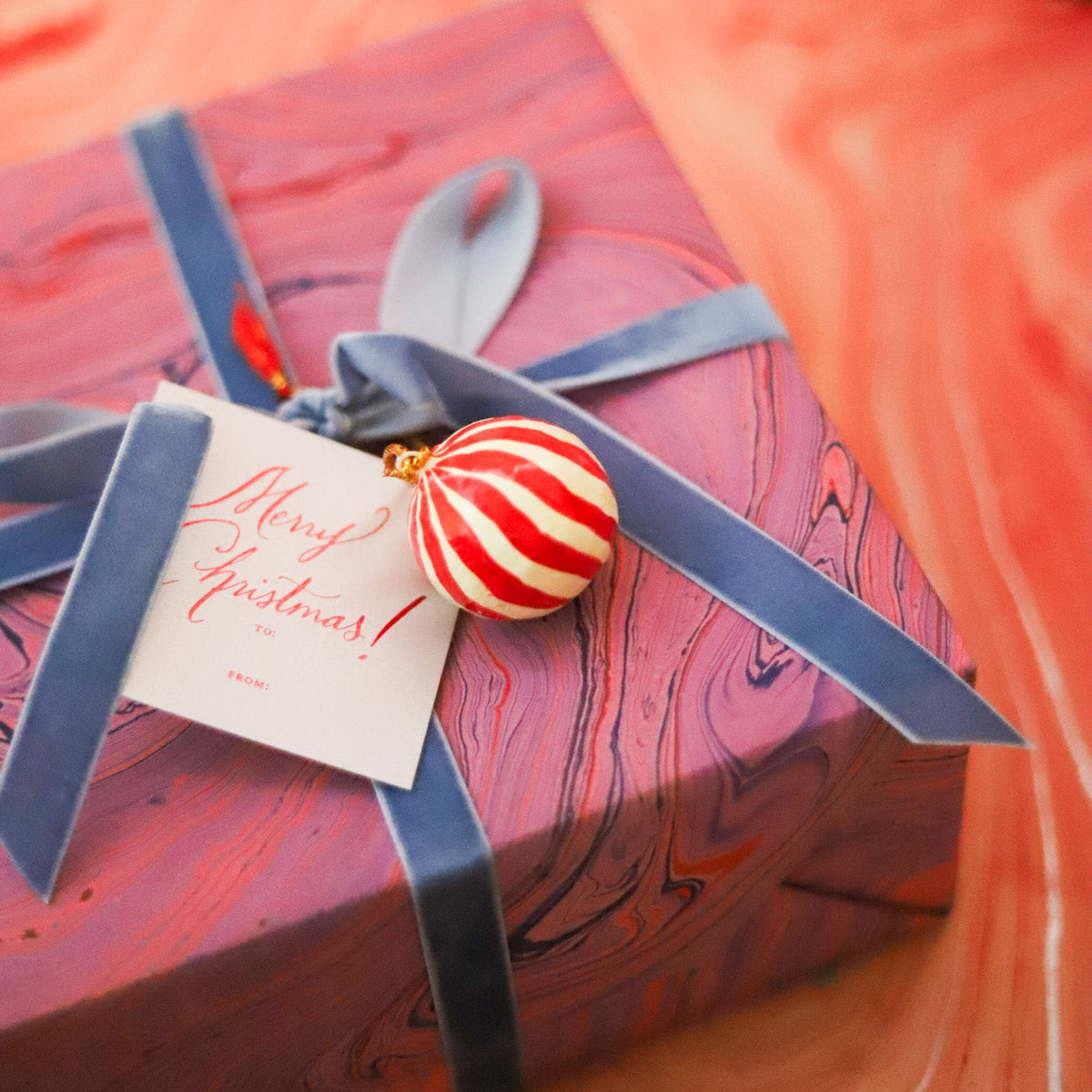 A wrapped gift box with a marbled pink and red pattern, tied with a blue ribbon and a small red-and-white striped ornament, with a card that reads 'Merry Christmas!'.