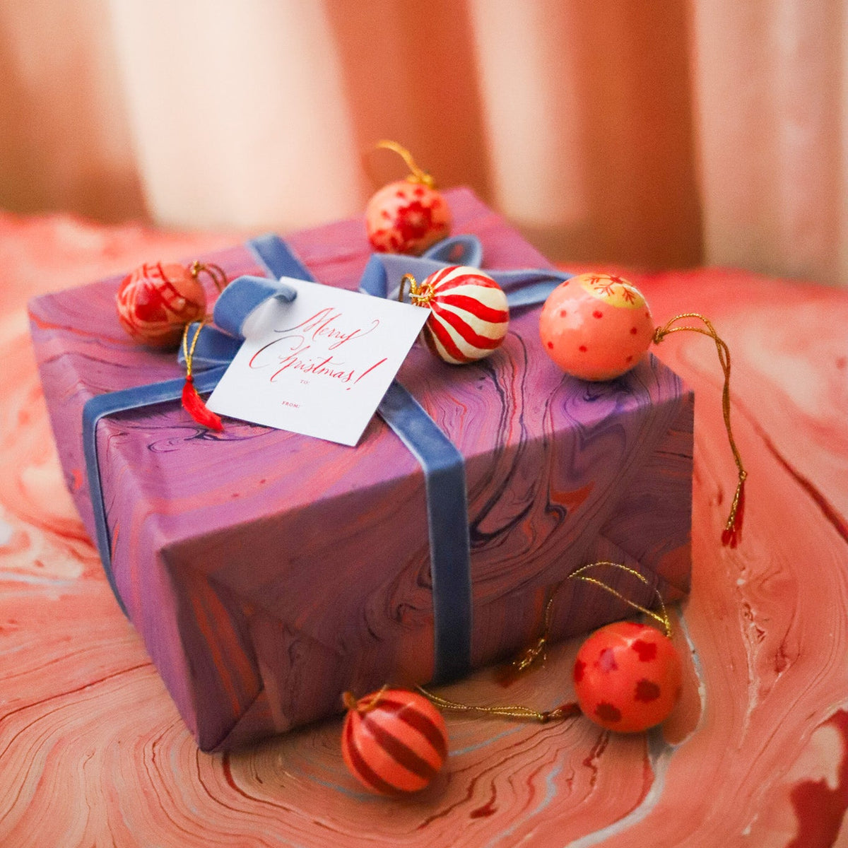 A wrapped gift box with purple marbled paper, a blue ribbon, and a white card reading 'Merry Christmas!' decorated with red and orange holiday ornaments, set on a pink marbled surface.