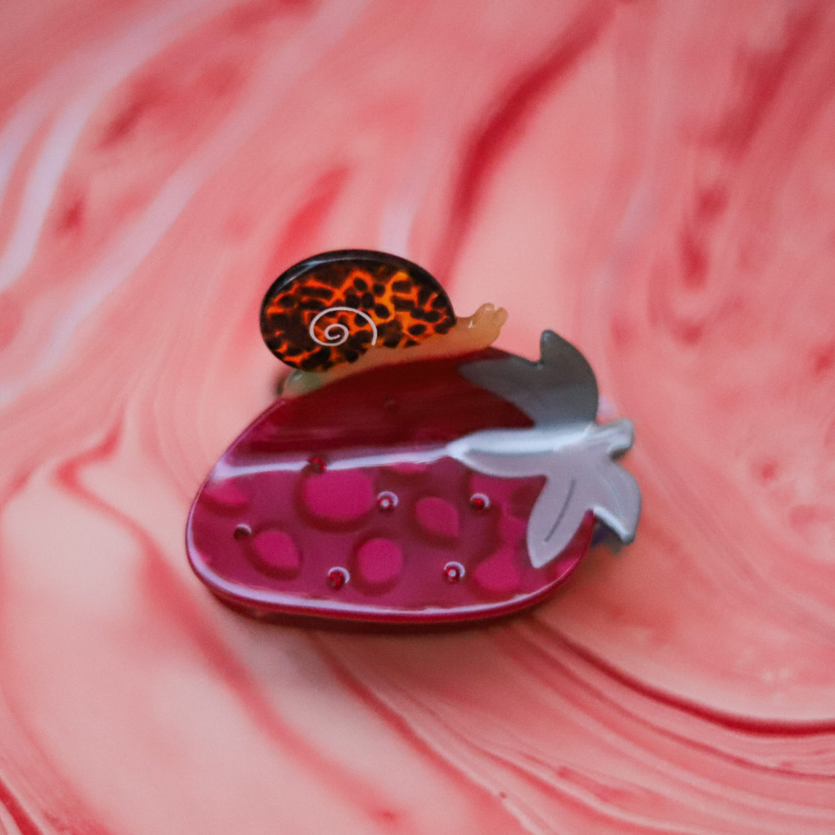 The Strawberry Snail Hair Claw features a glossy red strawberry with green leaves and a tiny tortoise shell snail, crafted from cellulose acetate.