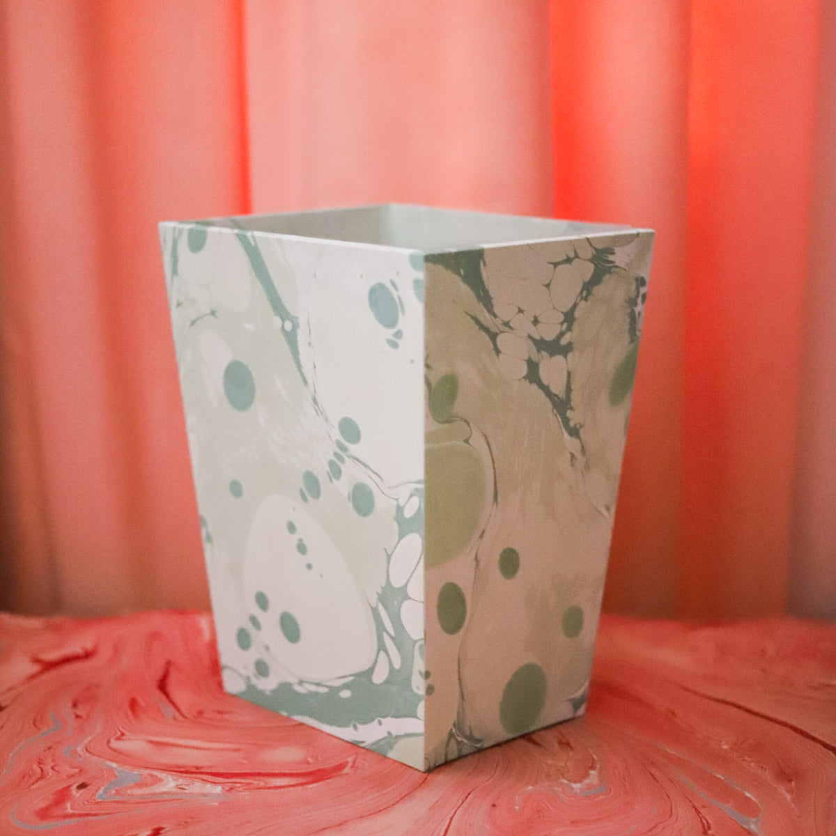 A square paper napkin with a green and beige marbled pattern and circular dot accents, standing upright on a textured pink surface with a pink background.