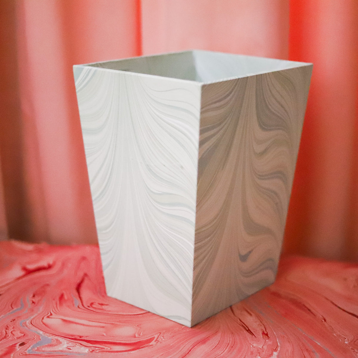 A square decorative planter with a white and beige marbled swirl pattern, set against a textured red background.