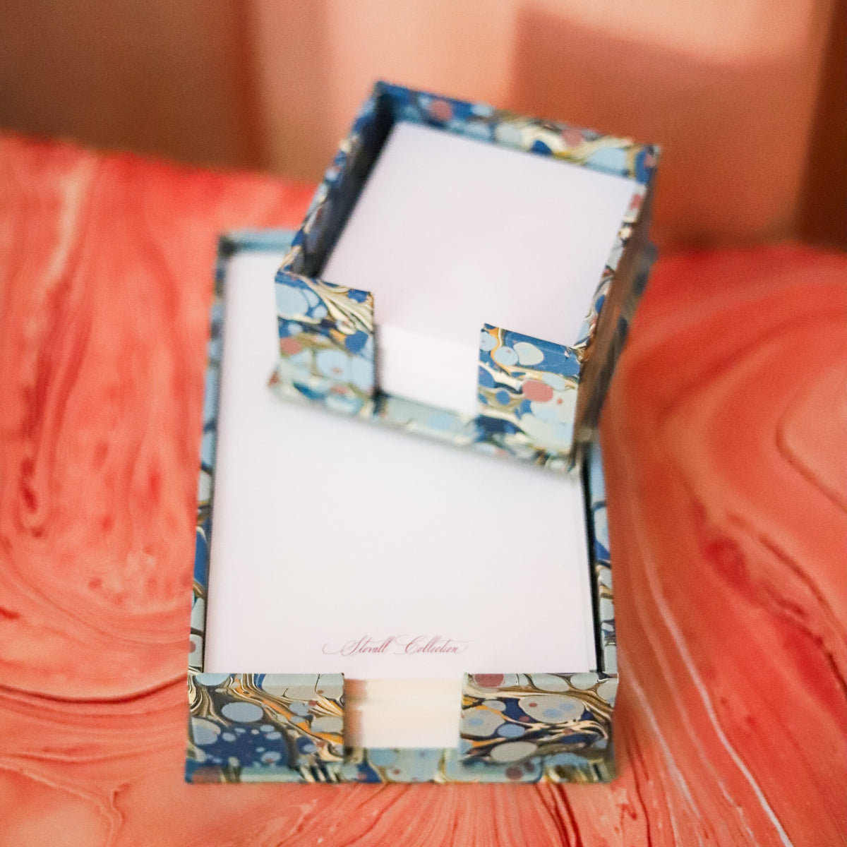 A decorative stationery set in a marbled blue, gold, and white patterned box, featuring a white notepad and matching envelope on a textured coral-pink surface.