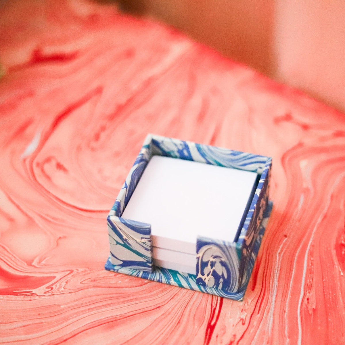 A small open jewelry box with a blue and white marbled pattern, containing white cards or tissue, placed on a coral-pink marbled surface.