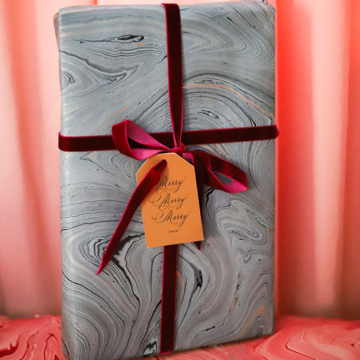 A wrapped rectangular gift box with a gray and white marbled pattern, tied with a dark red ribbon and bow, and adorned with a small orange gift tag, set against a pink marbled backdrop.
