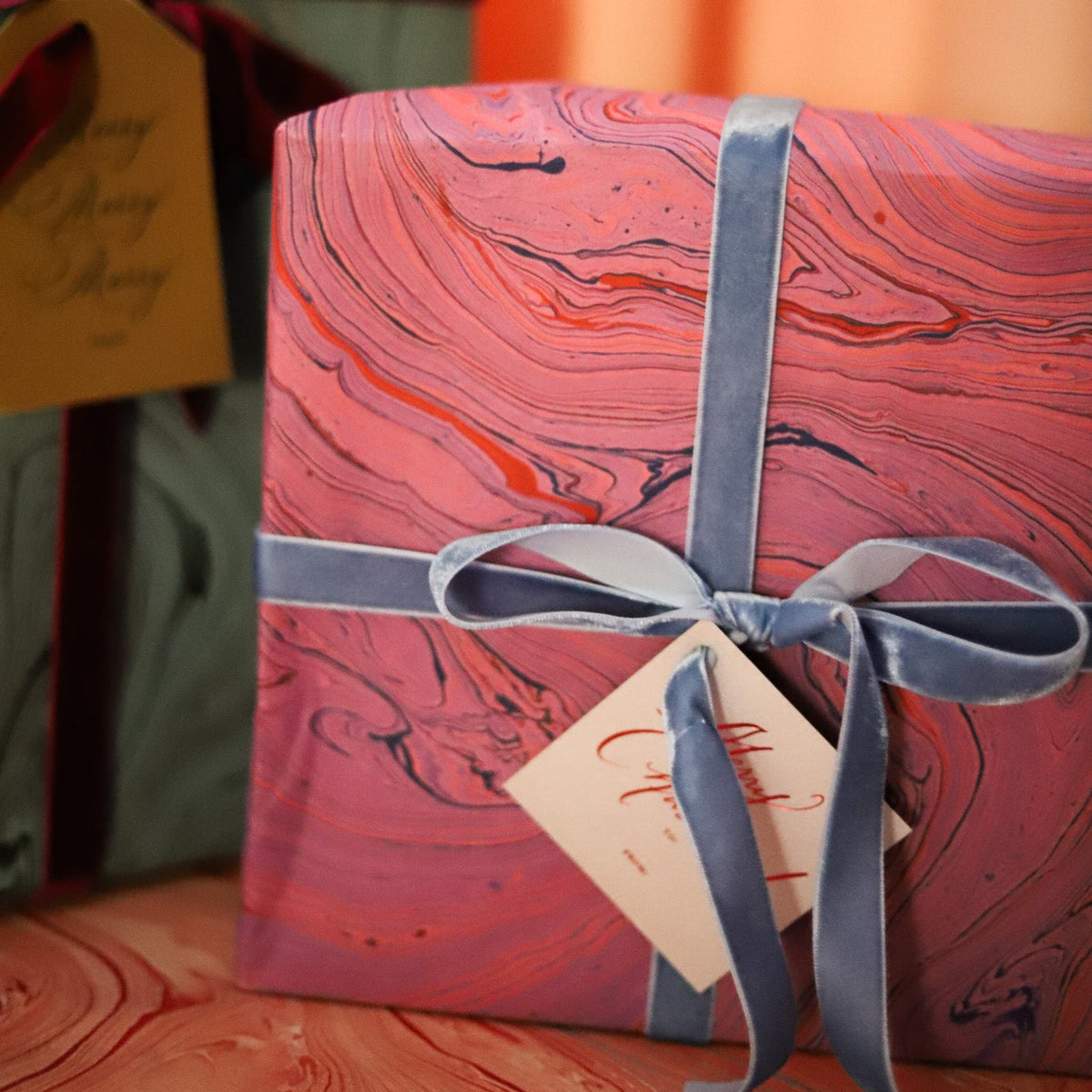A square gift box wrapped in pink and red marbled paper with a dusty blue ribbon tied in a bow, with a small cream gift tag attached. Another wrapped box is partially visible in the background on a wooden surface.